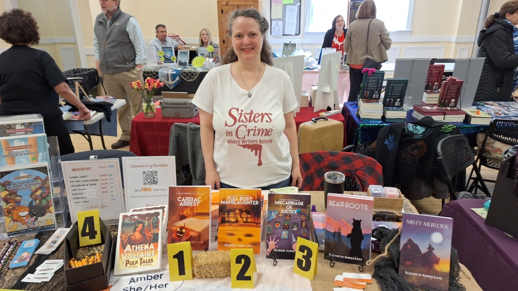 Amber wearing a Sisters in Crime t-shirt standing behind her author table
