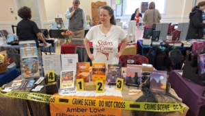 Amber wearing a Sisters in Crime t-shirt standing behind her author table