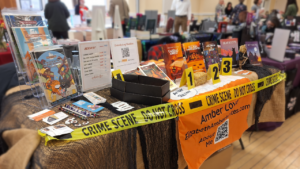 Amber's author table showing comics books on display and decorated like a creepy crime scene with yellow crime tape and evidence markers