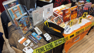 Amber's author table showing comics and books on display and decorated like a creepy crime scene with yellow crime tape and evidence markers