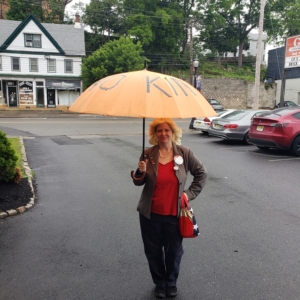 2025 #NoKings Day, Amber dressed as Leslie Knope from Parks and Recreation TV show holding orange umbrella with "No Kings" in black tape around it.