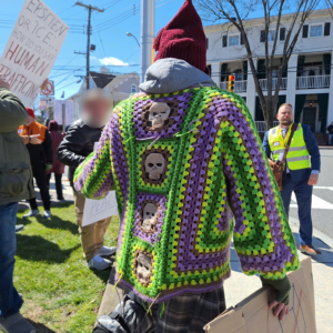 The back of a man who crochets wearing a colorful crocheted sweater with skulls down the back and the "melt ICE" hat based on Norway's red knit hats with tassels from WWII.