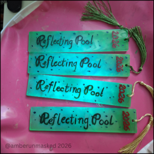 Under the glow of a blacklight, a silicone mat holding 4 resin art pieces: long neon green with speckled blue, hand-painted script, "Reflecting Pool" and "2026". Two have green tassels and the other two have gold tassels. The pieces have UV reactive glow.
